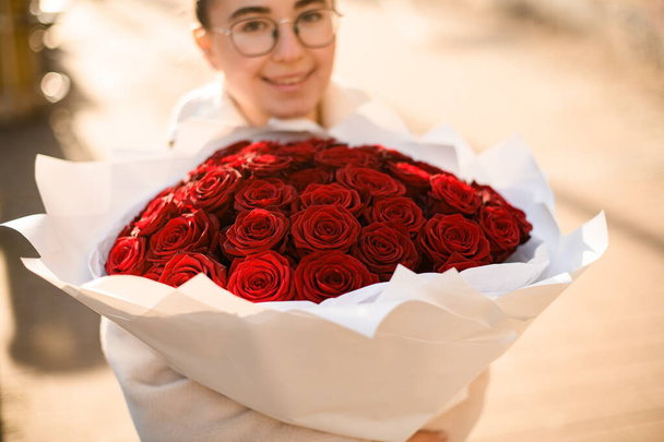 Big classy bright bouquet of red roses in hands of cute girl. One hundred and one roses in craft wrapping paper. Flowers shop concept - 写真・画像