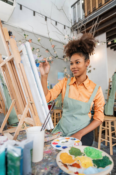 Νεαρή καλλιτέχνης όμορφη Αμερικανίδα Αφρικανή εργάζεται στο στούντιο ζωγραφικής της. - Φωτογραφία, εικόνα
