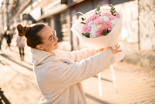 Young woman florist rising up freshly made minimalistic blossoming bouquet of pink, white carnations and eucalyptus branches formed in white wrapping paper on blurred city street background - Fotó, kép