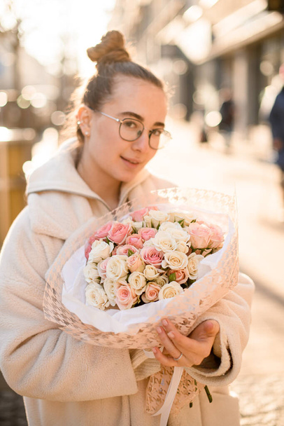Young attractive girl holding tender bouquet of small roses in pink and milk colors formed with wrapping paper and silk ribbon. Blurred background. Front view - Foto, Bild