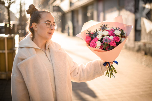 Young girl holds flowers bouquet with creamy and pink roses and thistles in wrapping paper and blue satin ribbon with flower shop logo. Bouquet in womans hands. Flower delivery - Foto, Imagem