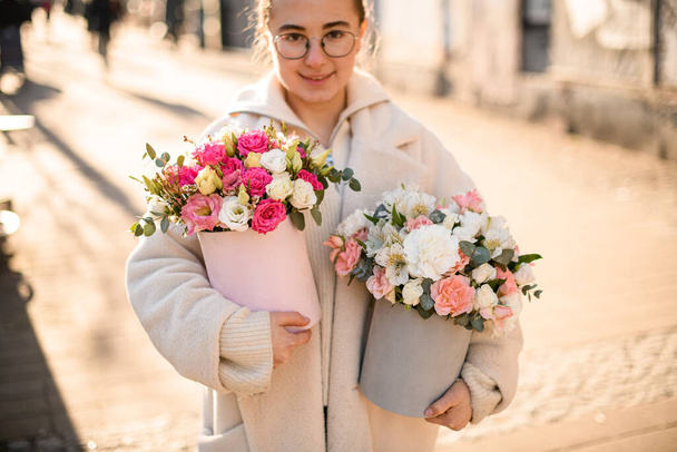 Cute girl with two lush flower bouquets with fresh roses, eustomas, alstroemerias and eucalyptus branches in hat boxes outdoors. Blurred background. Flower shop. Flower delivery - Foto, Imagem