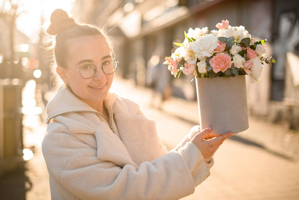 Young girl florist holding flower bouquet with fresh roses, alstroemerias and eucalyptus branches in gift box outdoors. Blurred background. Flower shop. Flower delivery - Foto, Imagen