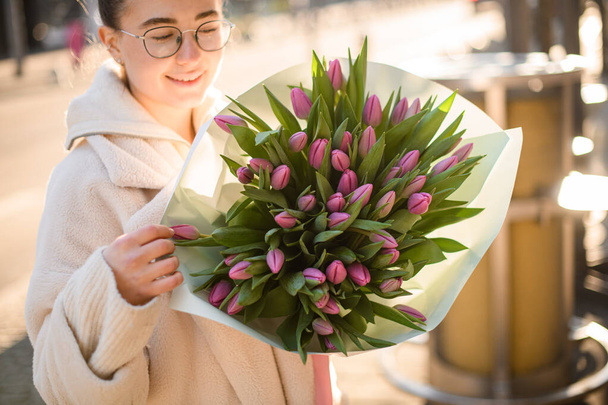 Young smiling woman holding delicate bouquet of pink fresh tulips in hands. Bunch of spring flowers in female hands - Fotó, kép