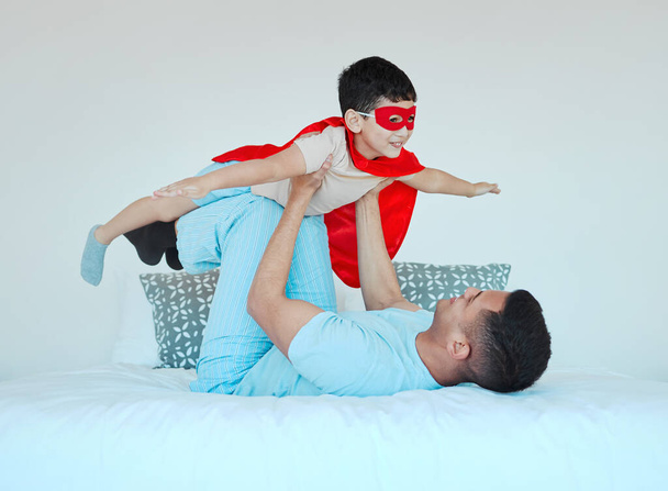 Samolot, sypialnia i tata bawią się z dzieckiem dla zabawy lub szczęścia w ich domu. Szczęśliwy, uśmiechnięty i młody ojciec relaksujący się ze swoim chłopcem w kostiumie superbohatera na łóżku w rodzinnym domu. - Zdjęcie, obraz