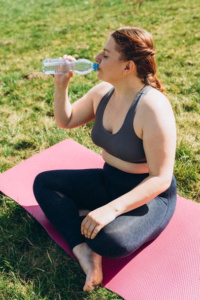 Attractive young 30s girl finished her work out and now drinking water in nature. She is sitting on a yoga mat on grass. - Фото, изображение