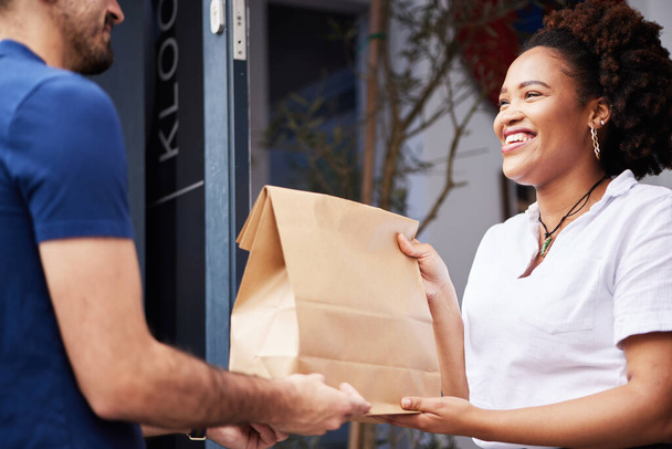 Παράδοση άνθρωπος, πακέτο και μια γυναίκα στην πόρτα με ένα χαμόγελο και χάρτινη τσάντα για το ηλεκτρονικό εμπόριο και τη ναυτιλία. Logistics, online αγορές και εμπορευμάτων ή ταχυμεταφορών εργαζόμενος δίνοντας μια ευτυχισμένη πελάτη μια παραγγελία fast food. - Φωτογραφία, εικόνα