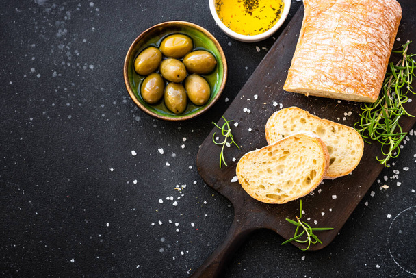 Ciabatta brood op houten plank met olijfolie, olijven en kruiden op zwart. Bovenaanzicht met kopieerruimte. - Foto, afbeelding