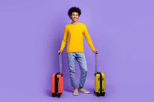 Full size photo of cheerful positive guy dressed yellow long sleeve jeans hold baggage in airport isolated on purple color background. - Foto, immagini
