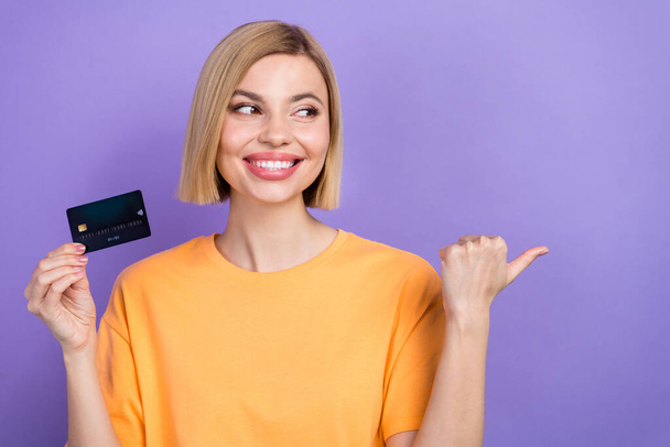 Portrait of cheerful girl dressed stylish clothes hold debit card look directing at offer empty space isolated on violet color background. - 写真・画像