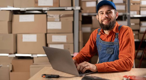 Ritratto di un giovane operaio felice che lavora al computer portatile in magazzino, preparando pacchi per la consegna. Copia spazio - Foto, immagini
