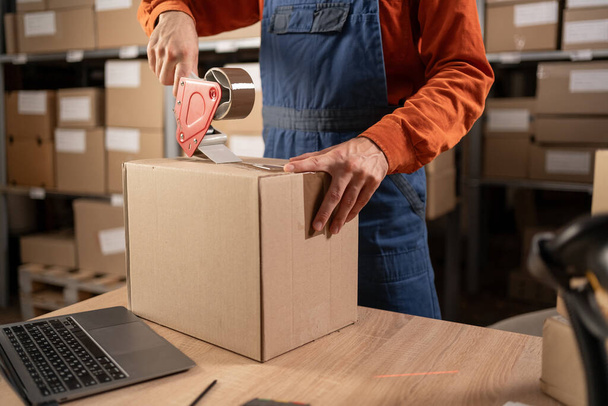 Worker man In Retail Warehouse working packing parcel box with tape dispenser for shipment. Delivery and distribution center full of shelves with goods. Close-up. Copy space - Photo, Image