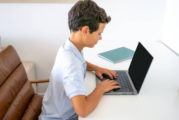Joyful teen guy writing at notebook and looking at laptop screen, having lesson online, home interior, free space - Fotoğraf, Görsel