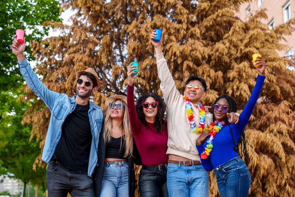 Grupo ecléctico de 5 amigos de diversos orígenes que celebran un alegre y colorido cumpleaños en el parque de la ciudad con bebidas, gafas de sol y risas bajo un árbol vibrante. - Foto, imagen