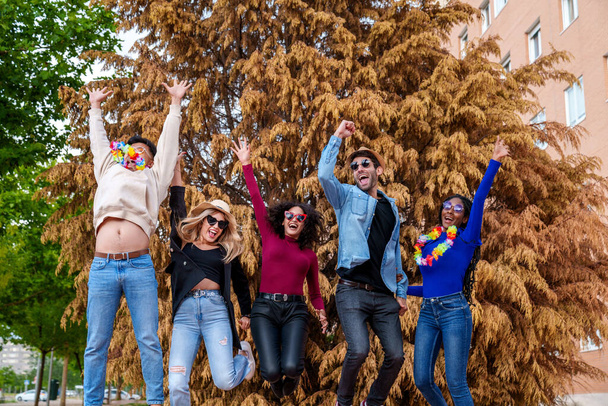 Ζωντανοί πολυεθνικοί φίλοι, στολισμένοι με εξοπλισμό για πάρτι, γιορτάζουν χαρούμενα σε ένα πάρκο της πόλης, δημιουργώντας μια πολύχρωμη και χαρούμενη ανάμνηση γενεθλίων. - Φωτογραφία, εικόνα