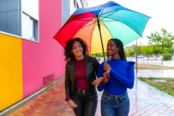 Regentag im Park: Zwei glückliche, vielfältige Freunde, ein Kaukasier und ein Afrikaner, halten einen LGBT-Regenschirm in der Hand und schaffen eine lebendige, bunte Szene. - Foto, Bild