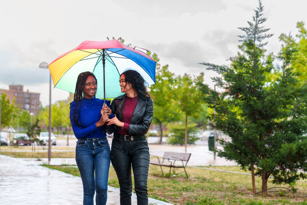 Δύο χαρούμενοι, ποικίλοι φίλοι, ένας Καυκάσιος και ένας Αφρικανός, αγκαλιάζονται κάτω από μια ζωντανή ομπρέλα ΛΟΑΤ, περπατώντας ευτυχισμένα ανάμεσα σε πολύχρωμα δέντρα σε ένα βροχερό πάρκο της πόλης. - Φωτογραφία, εικόνα
