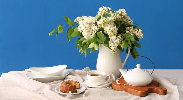 Bouquet of white lilac flowers, cup of coffee and tasty dessert on table near blue wall - Foto, Bild