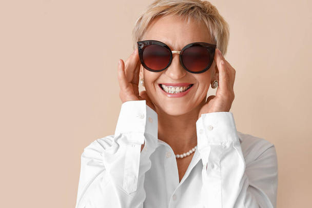 Beautiful mature woman in sunglasses on beige background, closeup - Photo, Image