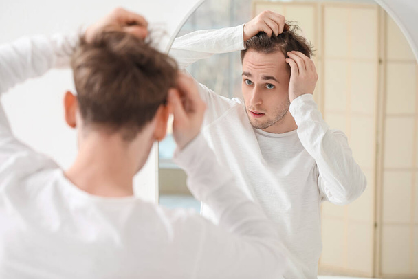 Молодой человек с проблемой выпадения волос смотрит в зеркало дома - Фото, изображение