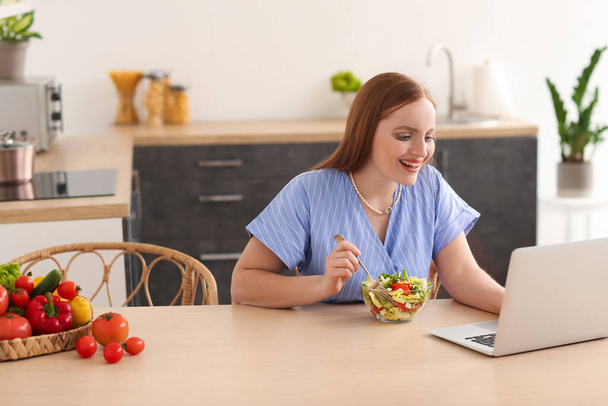 Young woman with laptop eating vegetable salad in kitchen - Photo, Image