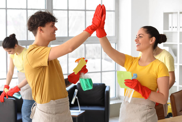 Jóvenes conserjes con suministros de limpieza que se dan chocan los cinco en la oficina - Foto, Imagen