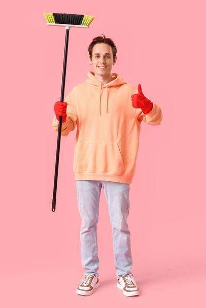 Young man with broom showing thumb-up on pink background - Fotografie, Obrázek