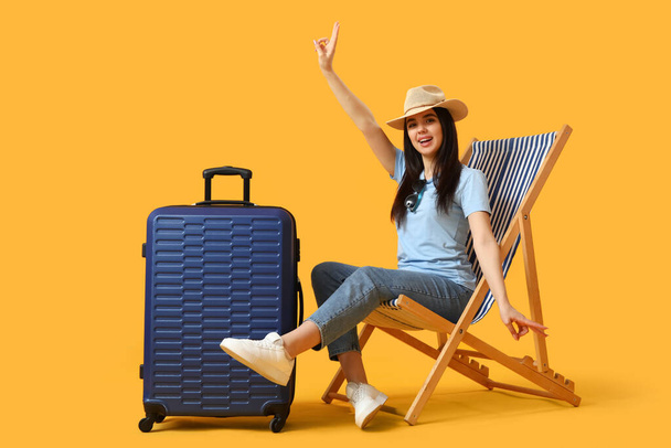 Young woman with suitcase in deck chair showing victory gesture on yellow background - Foto, Imagen