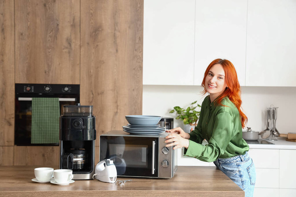 Joven pelirroja con cafetera, horno microondas y batidora en cocina moderna - Foto, Imagen