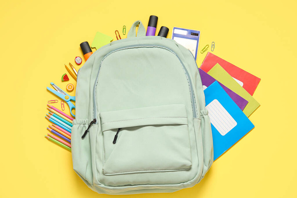 Πράσινη σχολική τσάντα πλάτης με διαφορετικά χαρτικά σε κίτρινο φόντο - Φωτογραφία, εικόνα
