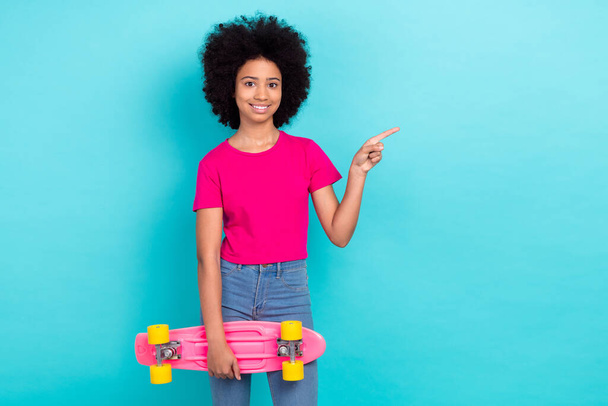 Photo of charming cool girl with afro chevelure wear pink t-shirt hold skateboard indicating empty space isolated on blue color background. - Photo, Image