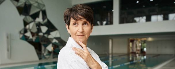 middle aged woman with short hair standing in white robe, spa center, indoors swimming pool, banner - Foto, Bild