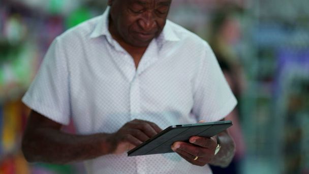 Un vieil homme brésilien noir joyeux utilisant un appareil tablette. Une personne afro-américaine plus âgée engagée dans la technologie moderne, regardant un écran souriant - Photo, image