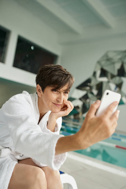 positive middle aged woman in white robe taking selfie, smartphone, spa center, swimming pool - Photo, Image