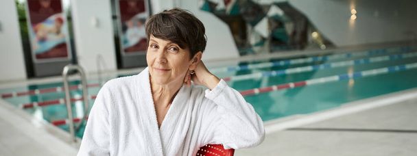 postura relaxada, mulher madura feliz em roupão branco sentado perto da piscina interior, dia de spa, banner - Foto, Imagem
