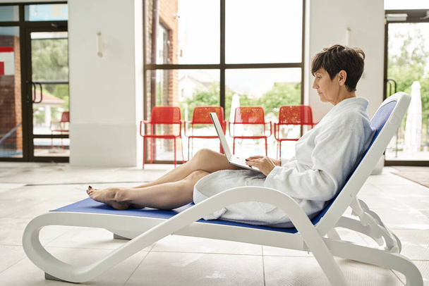 freischaffend und Wellness, Frau mittleren Alters im weißen Gewand mit Laptop, auf Liegen sitzend, Fernarbeit - Foto, Bild