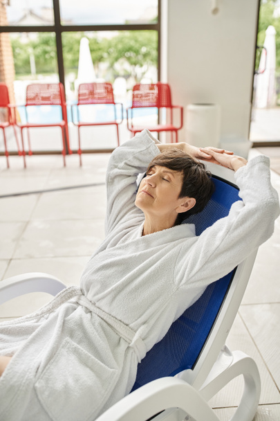 femme d'âge moyen avec les cheveux courts relaxant sur une chaise longue, robe blanche, centre spa, les yeux fermés - Photo, image