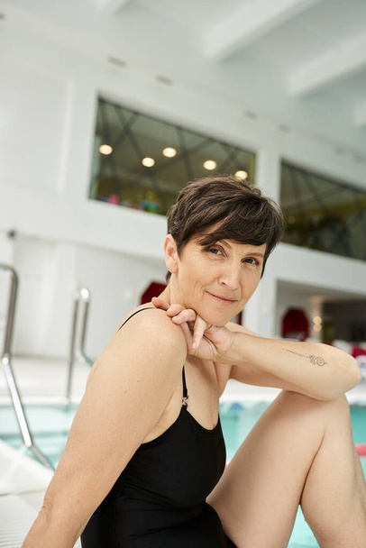 retrato de mujer de mediana edad tatuada en traje de baño sentado cerca de la piscina, junto a la piscina, centro de spa - Foto, imagen