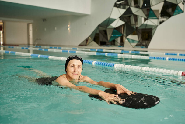 mujer de mediana edad nadando con tabla flotante en la piscina, gorra de natación y gafas, deporte, estilo de vida - Foto, Imagen