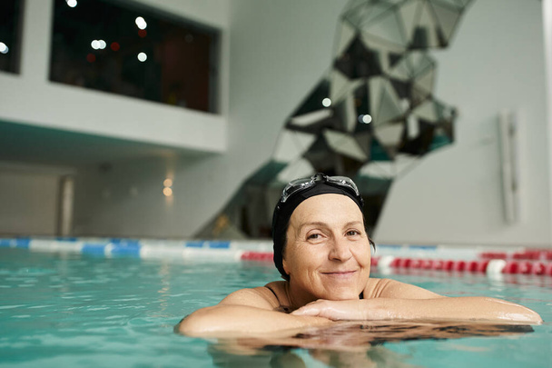 feliz mujer de mediana edad nadando con tabla flotante en la piscina, gorra de natación y gafas, sonrisa - Foto, Imagen