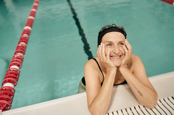 middle aged woman in swim cap and goggles looking away, smiling, water in pool, recreation center - Photo, Image