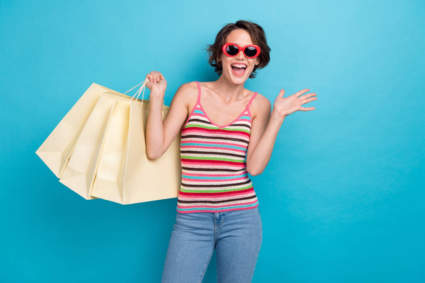 Photo of impressed funky woman dressed tank top dark eyewear holding shoppers isolated blue color background. - Photo, Image