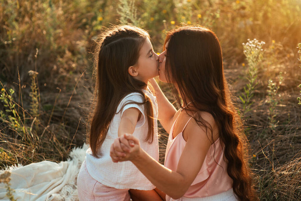 Hermosa madre joven y dulce hija joven abrazándose y besándose en una manta blanca entre un campo de flores al atardecer. Mamá y su hija pasar tiempo con feliz al aire libre - Foto, imagen