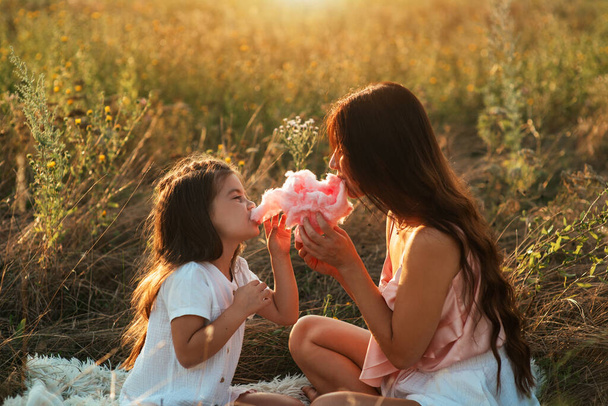 Красивая молодая мать и милая молодая дочь едят сахарную вату на белом одеяле среди поля цветов на закате. Мама и дочь проводят время на свежем воздухе - Фото, изображение