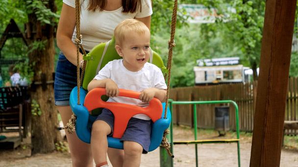 Щасливий хлопчик з матір'ю, яка гойдається на дитячому майданчику в літньому парку. Діти грають на відкритому повітрі, діти розважаються, літні канікули та свята
 - Фото, зображення