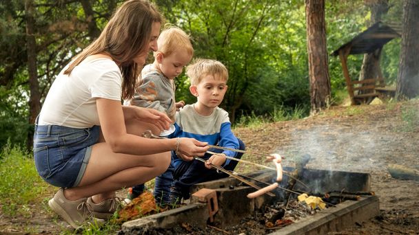 森のキャンプで燃える火の上でソーセージを調理する子供たちと幸せな家族。アクティブなレジャー、キャンプでの子供、自然の中での家族の休暇 - 写真・画像