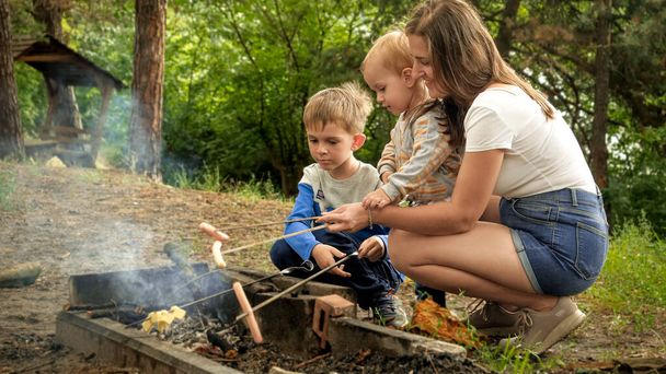 Jonge moeder met twee zoons die worstjes koken op kampvuur. Actieve recreatie, kinderen op de camping, familievakantie in de natuur - Foto, afbeelding