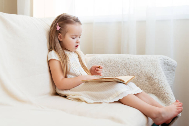 Pequeña chica rubia linda leyendo libro sentado en un sofá. Lectura infantil, ensueño y desarrollo de la imaginación. Día Internacional del Libro Infantil. A los niños les encanta leer. Día Mundial del Libro. - Foto, Imagen