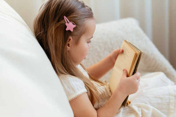 Klein schattig blond meisje dat een boek leest zittend op een bank. Kinderlezen, dromen en fantasieontwikkeling. Internationale Dag van het Kinderboek. Kinderen houden van lezen. Wereldboekendag. - Foto, afbeelding