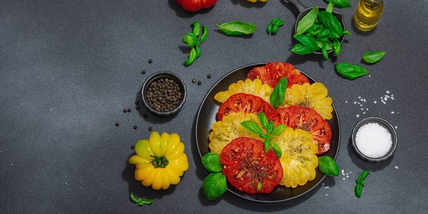 Colorful Heirloom tomato harvest. Ripe ribbed vegetables with fresh basil leaves. Juicy ingredient for summer salad, spices and oil. Hard light, dark shadow, black stone background, banner format - Photo, Image